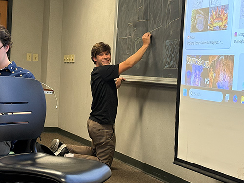 A student writing on a chalkboard and looking very pleased with himself at a TPE event