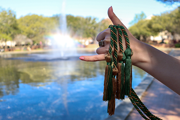 USC Graduation tassels held up by a hand making the 'spur' gesture with the Thomas Cooper library fountain in the background